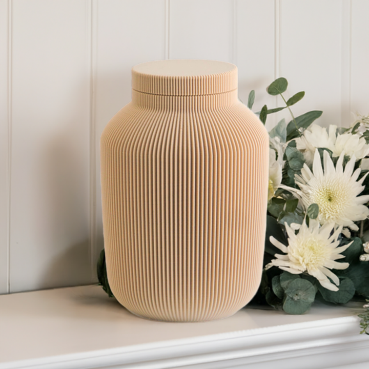 Willow cremation urn in tan with modern vertical sculpted texture, displayed with white flowers and greenery.