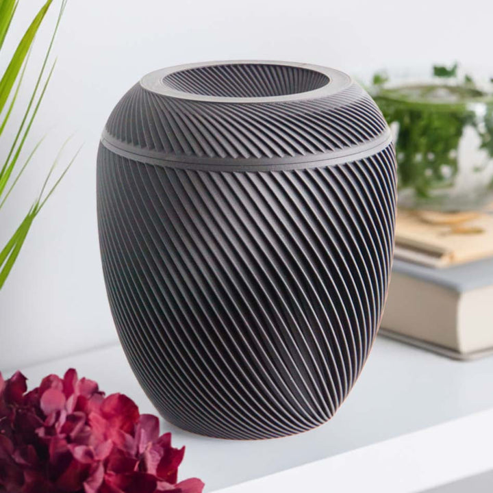 A matte black Laurel Urn with a spiral design, placed on a shelf with a plant and books in the background.