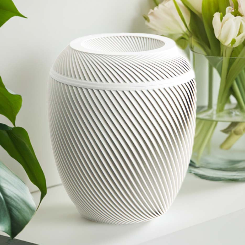 A white Laurel Urn with a spiral design, placed on a shelf with a plant and books in the background.