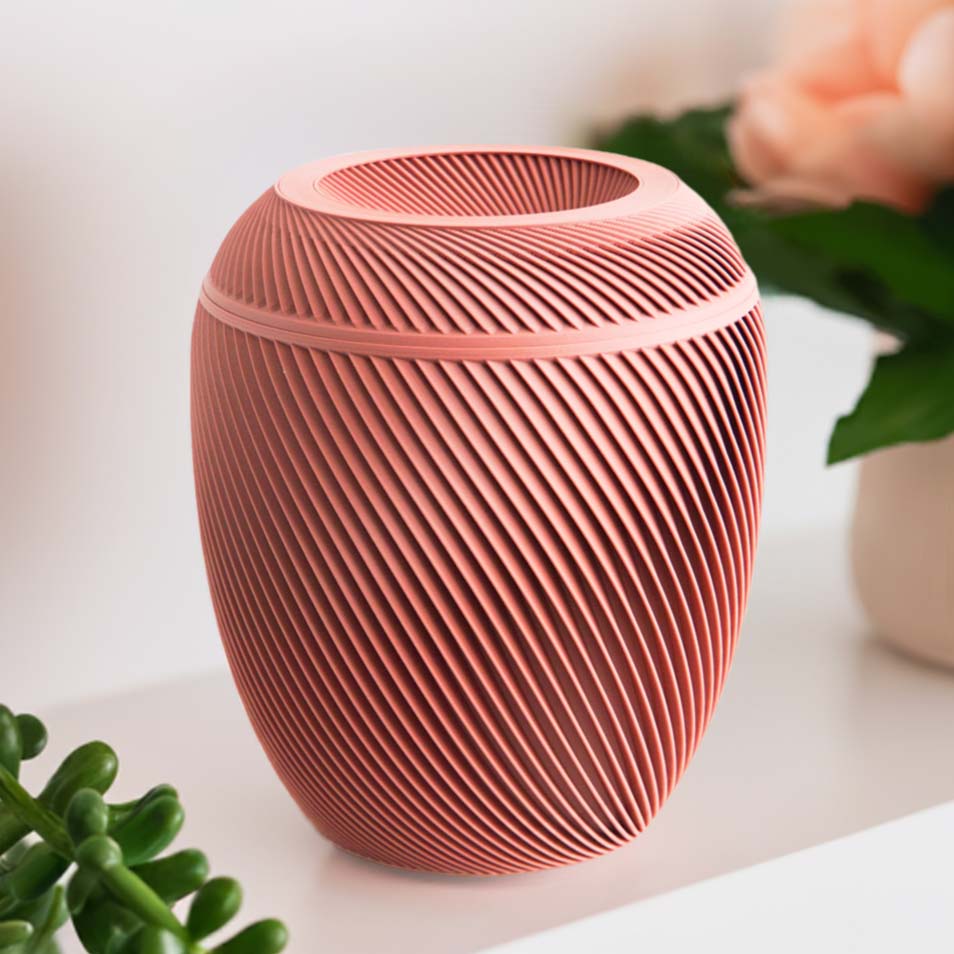 A salmon Laurel Urn with a spiral design, placed on a shelf with a plant and pink flowers in the background.
