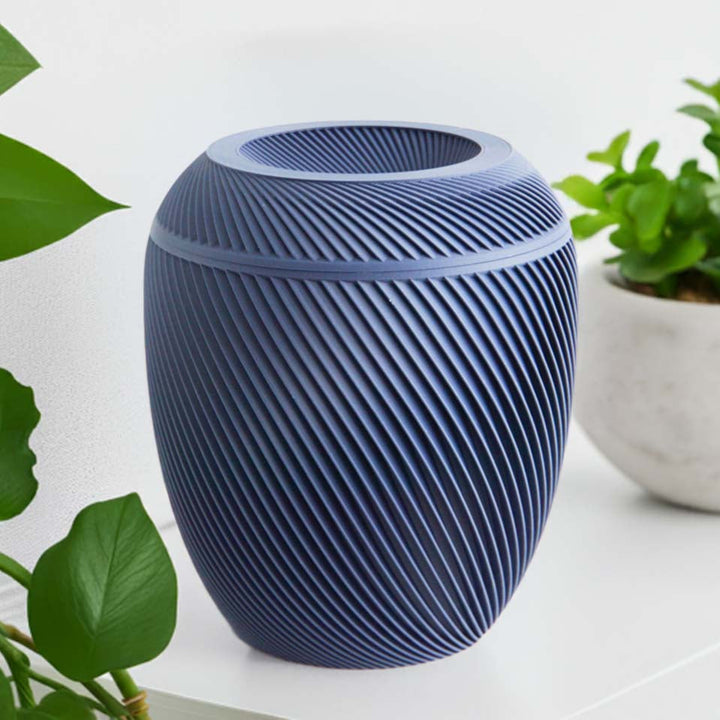 A navy Laurel Urn with a spiral design, placed on a shelf with plants in the background.